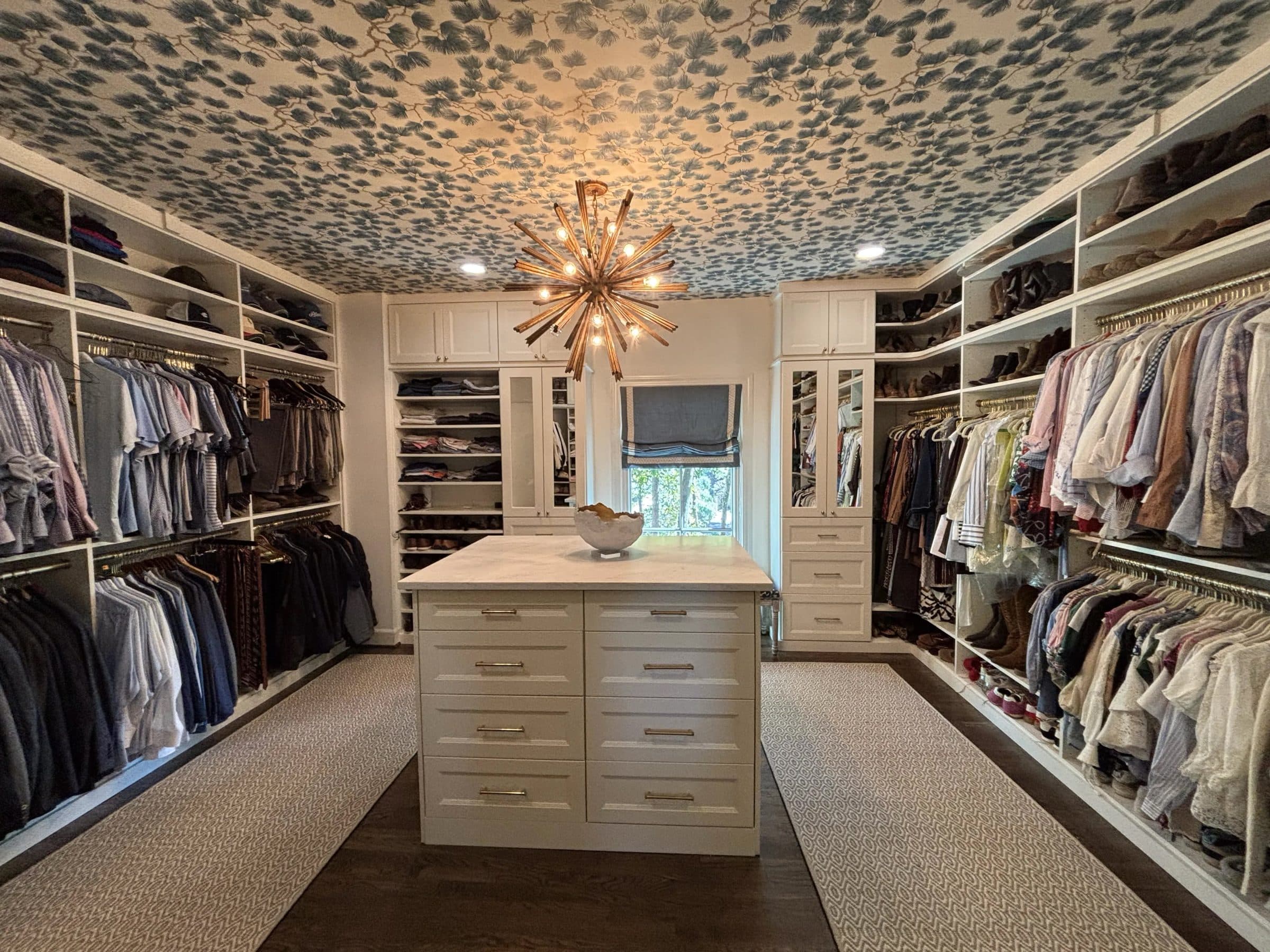 White walk-in closet with a center island, chandelier and wallpapered ceiling